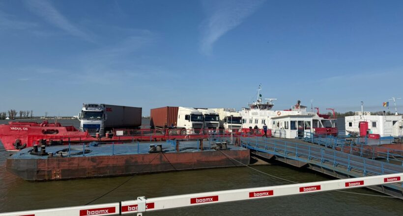 Port scene with a red oil barge, white trucks, and a pier with blue railings under a clear blue sky.