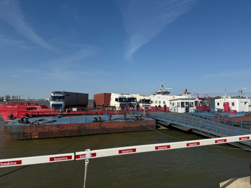 Port scene with a red oil barge, white trucks, and a pier with blue railings under a clear blue sky.
