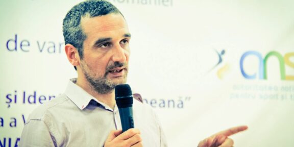 Man with short hair and beard speaks into a handheld microphone while gesturing with his right hand, standing in front of a Romanian event banner.
