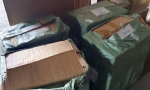 Stack of cardboard boxes wrapped in green plastic tarps, some taped and torn, stored in a corner.