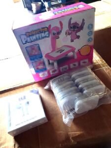 Pink kids' toy box with cartoon creatures and a pink activity table, beside a plastic-wrapped set of white discs.
