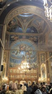 Inside ornate Orthodox church with a gilded iconostasis, frescoed arches, and a large chandelier above a dense congregation.