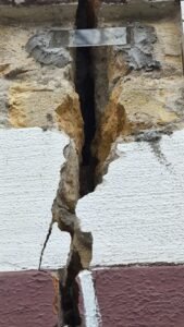 Cracked wall with a deep vertical fissure exposing rubble and a metal support beam at the top, indicating structural damage.