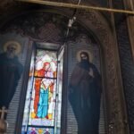 Interior of a church with a colorful stained-glass window of Mary and child, flanked by icons and ornate murals behind.
