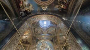 Ornate church ceiling with frescoes and arches, a blue circular window at the center, and a gold chandelier below; cracks run along the walls.