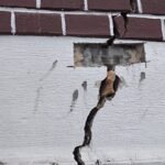 Exterior wall with a deep vertical crack running through brick and plaster, exposing inner material and rubble below a brick rowConsistency.