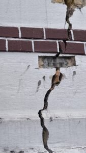 Exterior wall with a deep vertical crack running through brick and plaster, exposing inner material and rubble below a brick rowConsistency.