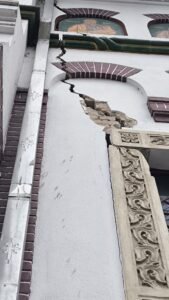 White building facade with a long vertical crack in the wall, exposed rubble, decorative carved stonework on the right, and a green pipe overhead.