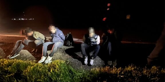 Five people in casual clothes sit on a rocky curb by a dark road at night, faces blurred, with backpacks nearby.