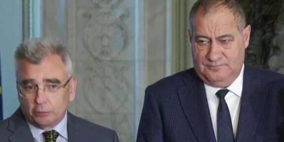 Two men in suits standing indoors during a press briefing, looking serious.