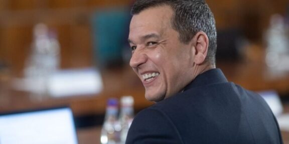 Man in a dark suit smiling over his shoulder in a conference room at a table with water bottles nearby
