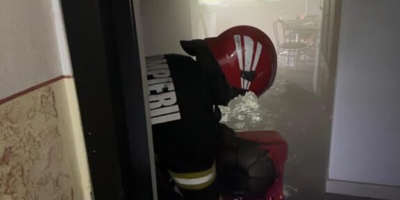 Firefighter in a red helmet and turnout gear crouches in a water-filled hallway during an emergency scene.