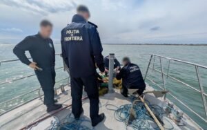 Border police officers on a boat work with fishing nets along a calm coastal waterway; their backs show 'Poliția de Frontieră' on uniforms (faces blurred).