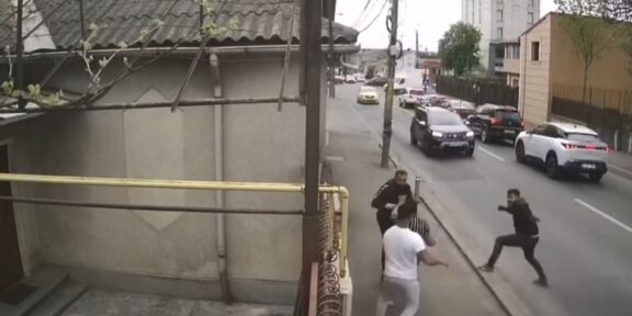 Group of men in a street confrontation on a sidewalk as cars drive by on a city road.