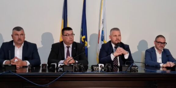Four men in suits sit at a press conference table with microphones, flags behind them.