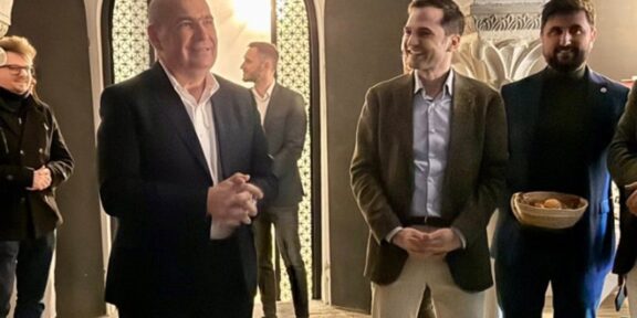 Group of people in a stone-arched hallway; a bald man in a dark suit speaks as a smiling man in a brown blazer stands nearby.