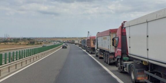 Row of large trucks lined up on the right shoulder of a highway, with a car in the left lane and open landscape beside the road.