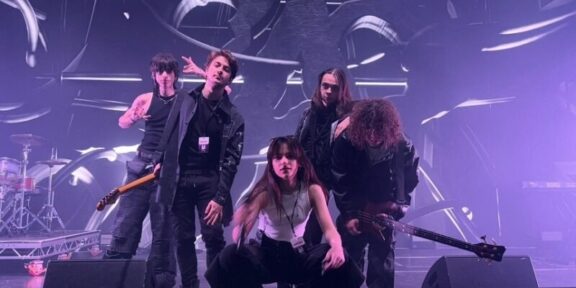 Five-person rock band posing on a stage with drums and guitars, under purple stage lights and abstract backdrop.