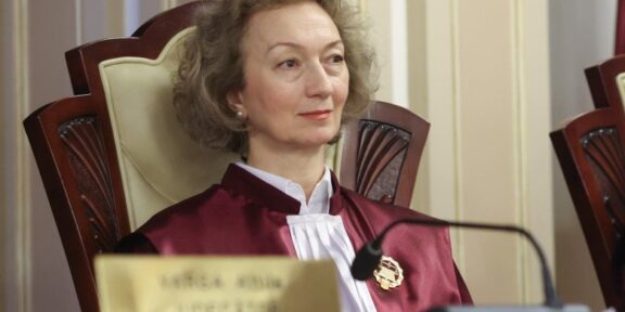 Female judge in a burgundy robe sits at a courtroom bench, looking slight to the right, with a nameplate and microphone in front of her.