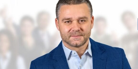 Professional portrait of a man in a blue blazer, foreground, with a crowd blurred in the background.