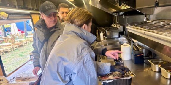 Inside a stainless steel food truck kitchen, a woman in a blue jacket places food from a foil pan while two men watch from behind.
