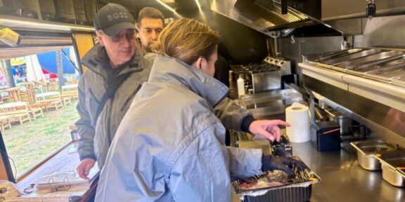 Inside a stainless steel food truck kitchen, a woman in a blue jacket places food from a foil pan while two men watch from behind.