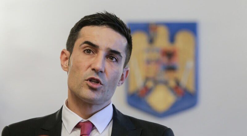 Man in a dark suit and red tie speaking at a press conference, with a blurred emblem in the background.