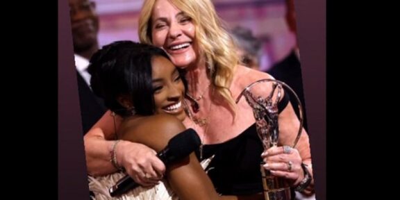 Two women hug on stage at an awards ceremony, smiling, with a crystal trophy visible.