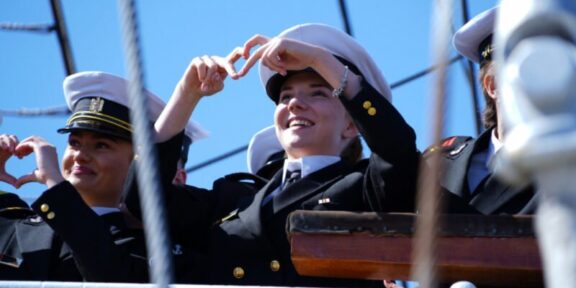 Cadets in navy uniforms on a ship deck making heart shapes with their hands against a clear blue sky.