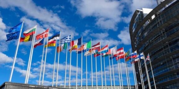 Row of colorful national flags on tall poles in front of a modern curved building against a blue sky outside an office complex.