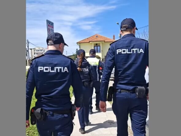 Group of police officers in dark blue uniforms labeled POLITIA walking along a sidewalk past a yellow house.
