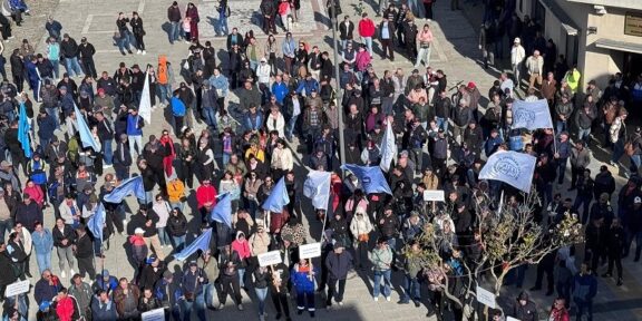 Aerial view of a large crowd in a city square participating in a protest, holding blue flags and white placards.