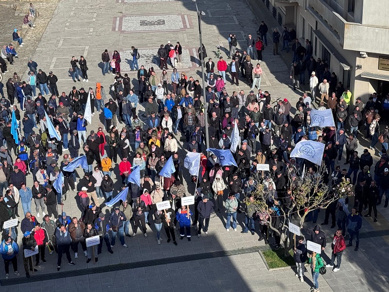 Aerial view of a large crowd in a city square participating in a protest, holding blue flags and white placards.