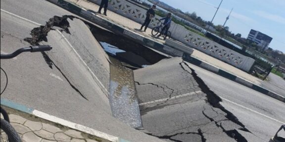 A wide street sinkhole with cracked asphalt, filled with water, spilling toward the curb while three people and a cyclist observe from the sidewalk behind a decorative railing.