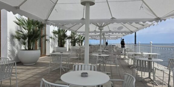Outdoor seaside cafe with white tables and chairs under large white shade umbrellas along a railing overlooking the ocean.