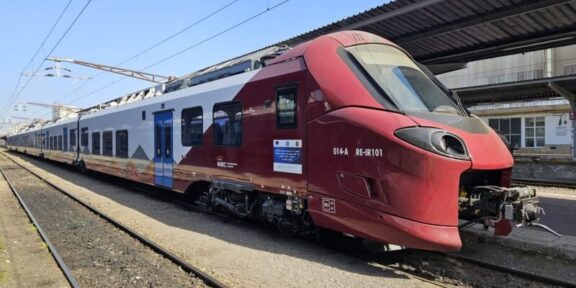 Red and white passenger train parked at a platform with overhead electric lines visible.