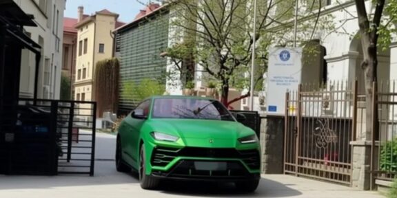 Bright green Lamborghini SUV driving down a gated residential street with parked cars and trees on a sunny day.