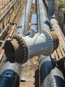 Large gray metal pipelines with flanged joints in a muddy trench at a construction site.