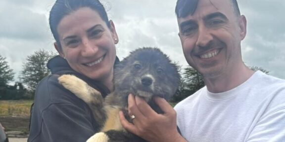 Smiling couple holding a fluffy puppy outdoors against a cloudy skyp