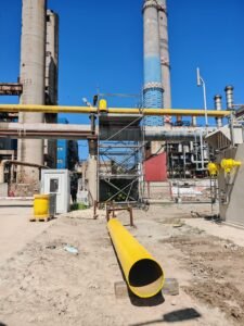 Industrial plant with tall smokestacks and a bright yellow pipeline running overhead against a blue sky