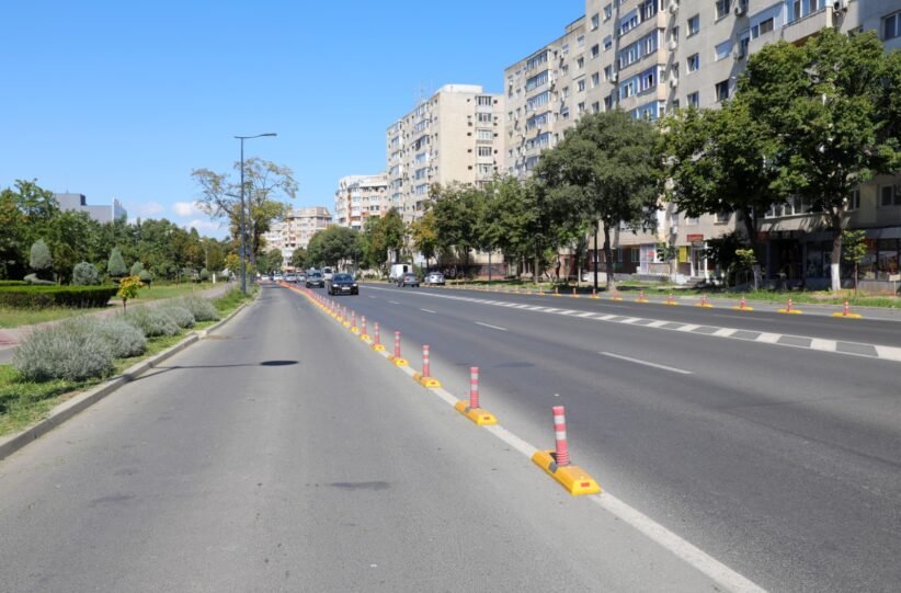 Wide city street with a central row of orange-and-white traffic posts and a sidewalk on the left, tall apartment buildings on the right.