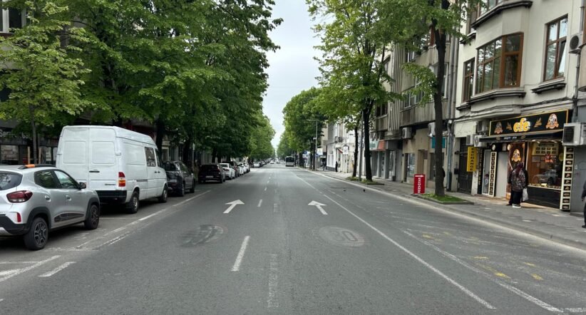 Urban street view with parked cars on both sides, green trees, and a shopfront on the right sidewalk aisle ahead in the distance traffic arrows on the road calm and overcast sky behind trees and buildings.