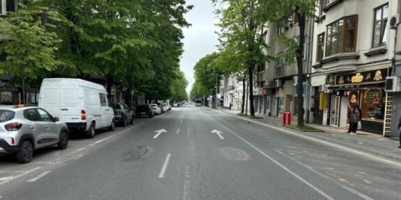 Urban street view with parked cars on both sides, green trees, and a shopfront on the right sidewalk aisle ahead in the distance traffic arrows on the road calm and overcast sky behind trees and buildings.