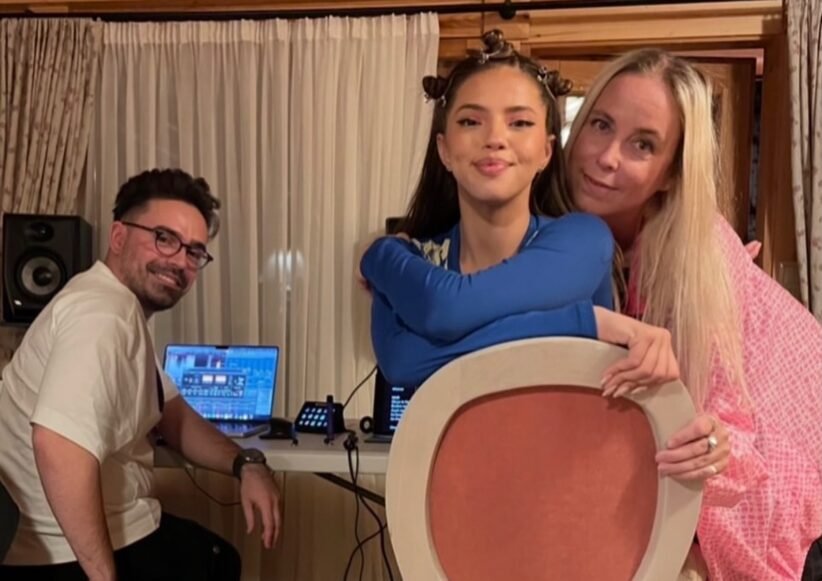 Three people pose for a photo indoors: a man with DJ equipment sits at a table with laptops, while a young woman in blue holds a large circular prop and an older woman in pink stands beside her.