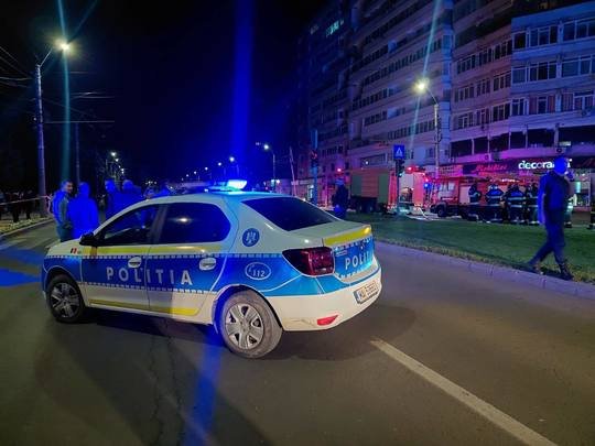 Blue-and-white Romanian police car with flashing blue lights on a nighttime city street, police and bystanders in the background.