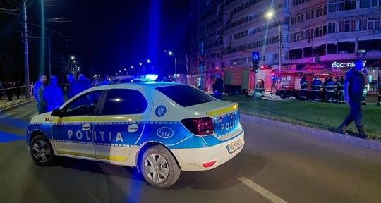 Romanian police car with blue lights on a nighttime city street; bystanders and a lit storefront in the background.