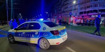 Romanian police car with blue lights on a nighttime city street; bystanders and a lit storefront in the background.