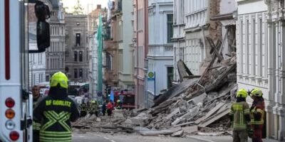 Firefighters in yellow helmets and reflective jackets assess a street blocked by debris from a collapsed building.