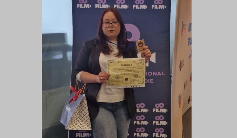 Woman with glasses in a blazer and white top holds a certificate and a trophy in front of a blue film festival banner, with a gift bag at her side.