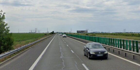 Black sedan in the right lane of a divided highway with green guardrails and open fields on both sides under a cloudy sky.
